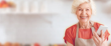 Smiling elderly woman in a red top and apron holding a smartphone in a bright, softly blurred kitchen
background.
