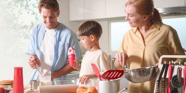 Happy family cooking together in a bright kitchen, with a child holding a red Kitchen Mama can opener, surrounded by Kitchen Mama tools like a gravity grinder, spatula, and whisk.
