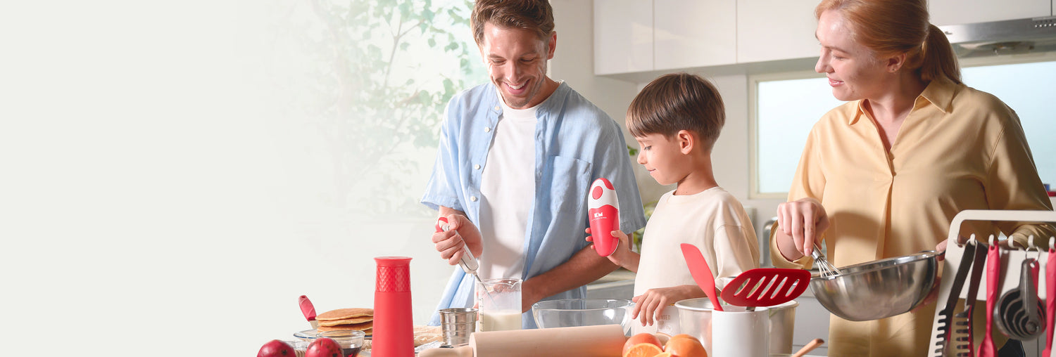 Happy family cooking together in a bright kitchen, with a child holding a red Kitchen Mama can opener, surrounded by Kitchen Mama tools like a gravity grinder, spatula, and whisk.