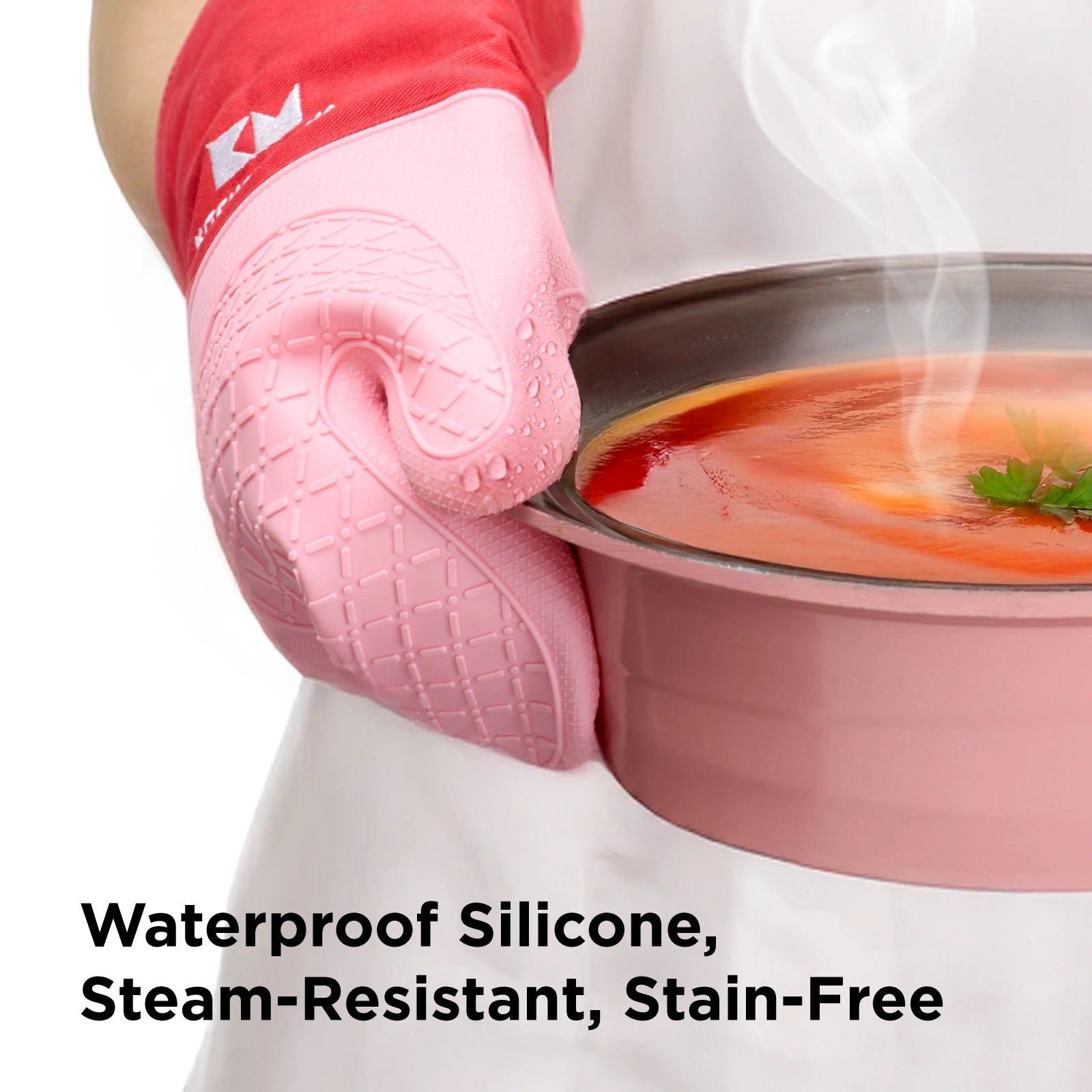 Close-up of a person holding a hot pot of tomato soup with red and pink Kitchen Mama silicone oven mitts. The mitts show water droplets on the surface, highlighting their waterproof, steam-resistant, and stain-free features. Text overlay reads: 